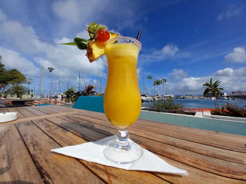 Cocktail exotique à base d'ananas et de mangue au premier plan. Vue sur les bateaux au port Moselle et le ciel bleu à l'arrière plan.
bars et restaurants à Nouméa