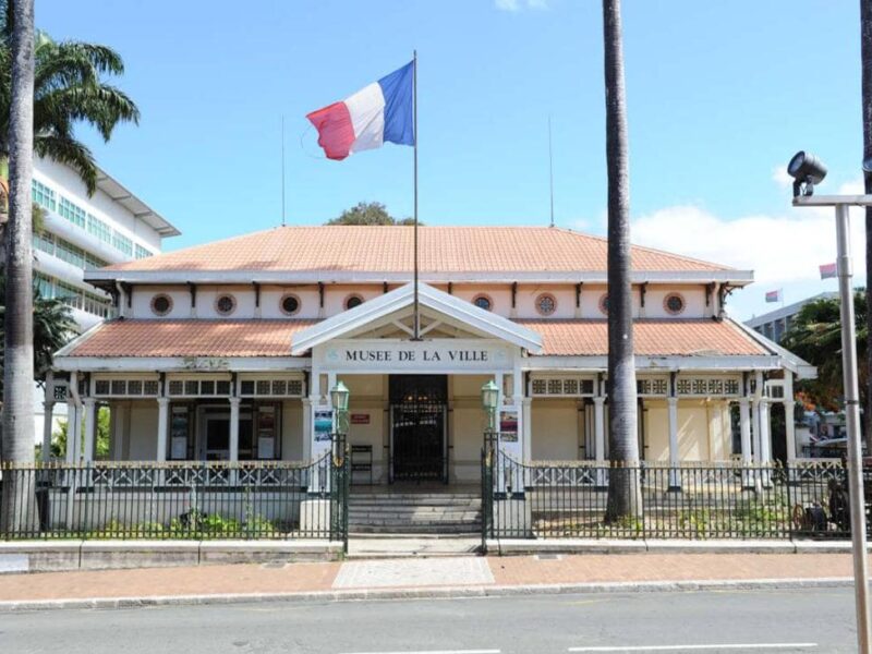 Photo du musée de la Ville de Nouméa vu de l'extérieur. Un drapeau français plane dans le ciel au-dessus du bâtiment. 