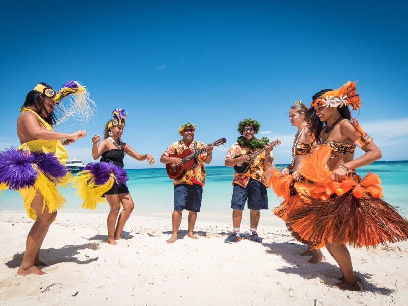 Photo de 4 danseuses tahitiennes et de 2 musiciens (jouant de la guitare et du ukulélé) sur la plage de l'îlot du Phare Amédée en Nouvelle-Calédonie.