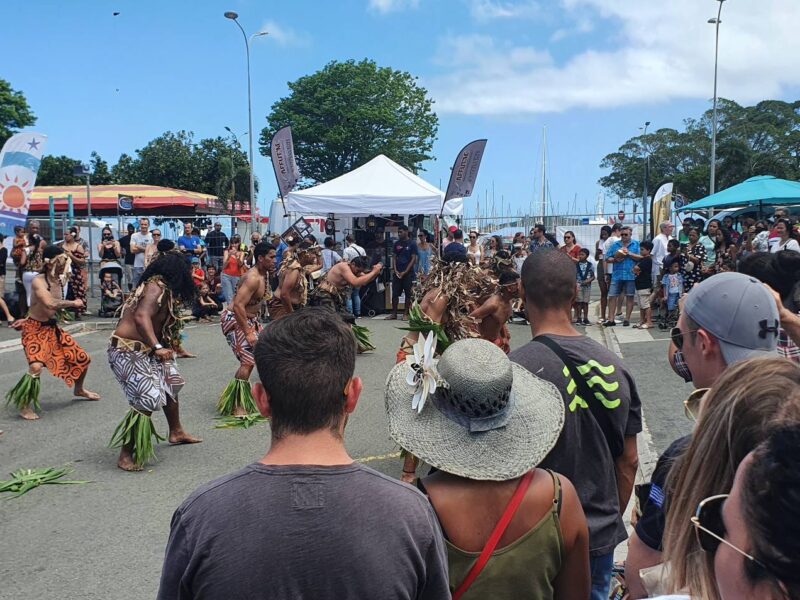Photo prise à la Foire du Pacifique en Nouvelle-Calédonie, lors d'un spectacle de danse kanak. On y voit plusieurs personnes regroupées autour d'une dizaine de danseurs vêtu de tenue traditionnelle kanak faisant un pilou.