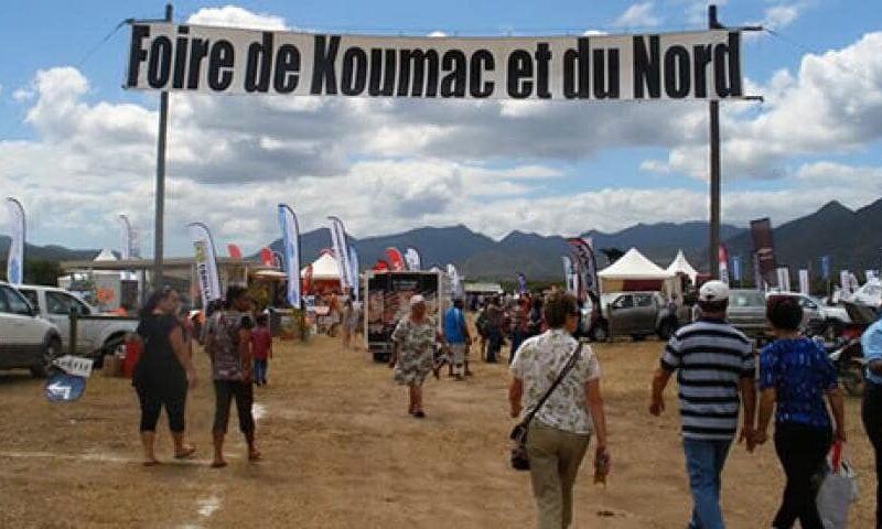 Photo de l'entrée de la Foire de Koumac en Nouvelle-Calédonie. On voit une banderole indiquant "Foire de Koumac et du Nord", des personnes y entrant ainsi que plusieurs stand en plein air.