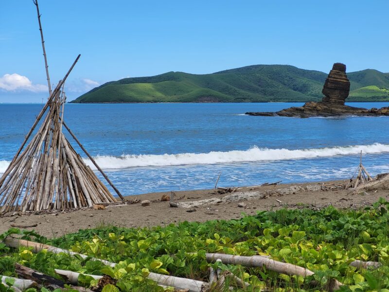 Photo d'un tipi en bois construit avec des bambous échoués sur la plage. Le bonhomme de Bourail est visible en arrière plan.